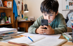 Criança concentrada no celular enquanto ignora livros, cadernos e materiais de estudo sobre a mesa