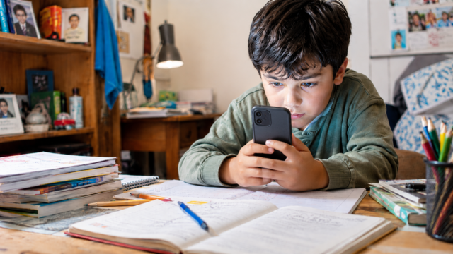 Criança concentrada no celular enquanto ignora livros, cadernos e materiais de estudo sobre a mesa