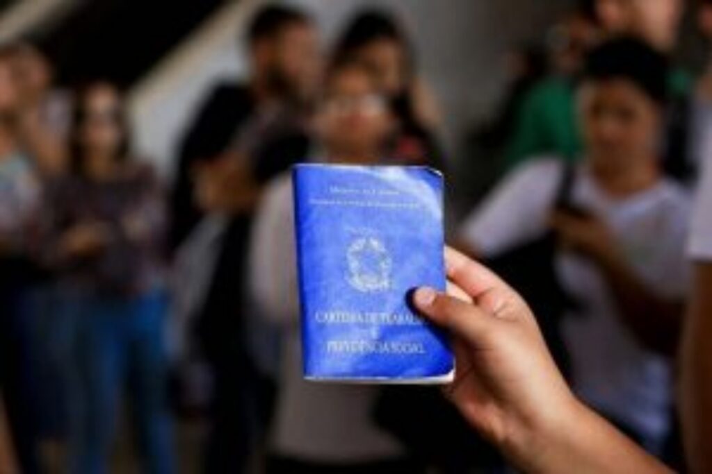 Pessoa segura uma carteira de trabalho diante de trabalhadores